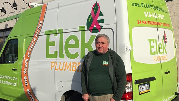  Technician in green shirt, standing in front of green and white Elek Plumbing service van.
